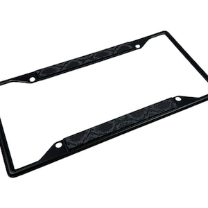 May include: A black license plate frame with a snakeskin pattern on the top and bottom. The frame is rectangular with rounded corners and has pre-drilled holes for mounting. The snakeskin pattern is a dark gray and black.
