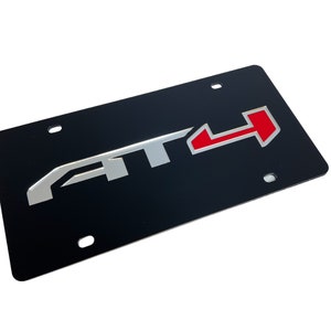 May include: Black license plate with a silver and red "AT4" logo. The logo is raised and has a 3D effect.