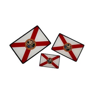 May include: Three framed Florida state flags of different sizes. The flags are white with a red diagonal cross and a state seal in the center.