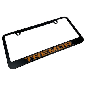 May include: Black license plate frame with the word "TREMOR" in orange lettering. The frame has a rectangular shape with rounded corners and mounting holes at the top. The lettering is in a bold, sans-serif font.