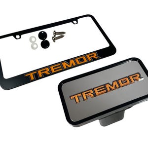 May include: Black license plate frame and a silver-colored hitch cover, both with the word "TREMOR" in orange lettering. Includes mounting hardware: screws, washers, and black caps. Automotive accessory.