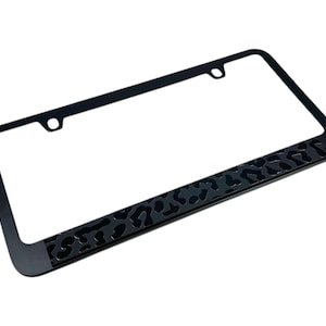 May include: Black metal license plate frame with a leopard print design on the bottom edge.