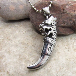 Puede incluir: Un collar con un colgante de plata con un diseño detallado y ornamentado de una cabeza y garra de dragón. El colgante está unido a una cadena de bolas de plata.