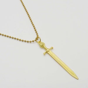 Pode incluir: Um colar de corrente de ouro com um pingente de espada. A espada tem uma empunhadura detalhada e uma l&acirc;mina longa e reta.