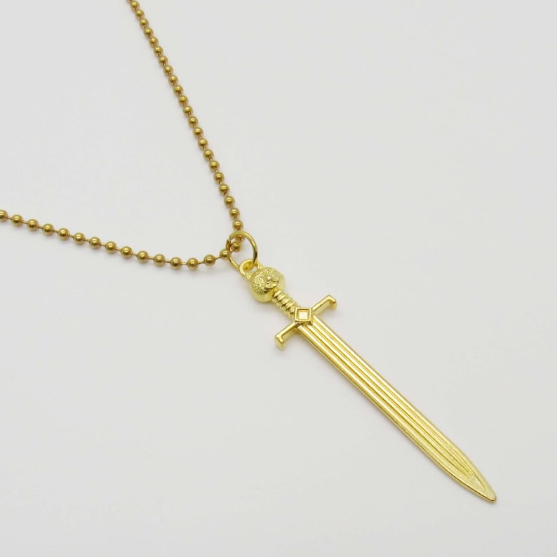Pode incluir: Um colar de corrente de ouro com um pingente de espada. A espada tem uma empunhadura detalhada e uma l&acirc;mina longa e reta.