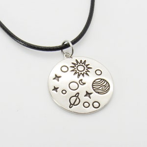 May include: A silver necklace with a round pendant featuring a black and white illustration of the sun, moon, stars, and planets. The necklace is on a black cord.