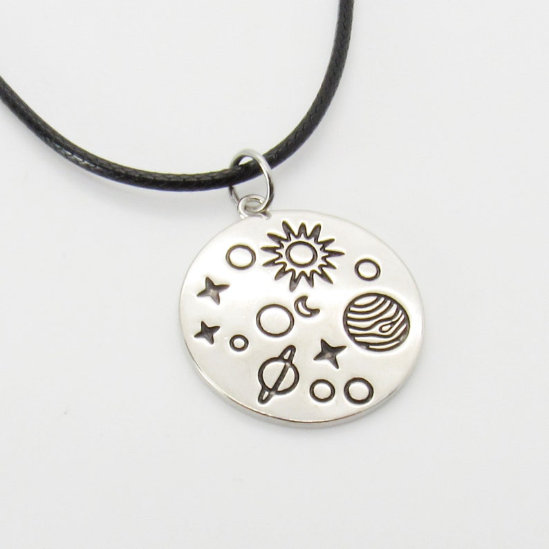 May include: A silver necklace with a round pendant featuring a black and white illustration of the sun, moon, stars, and planets. The necklace is on a black cord.