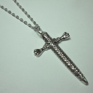 May include: A silver chain necklace with a sword-shaped cross pendant. The cross is detailed with a braided design and has a pointed tip.