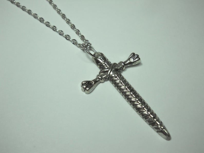 May include: A silver chain necklace with a sword-shaped cross pendant. The cross is detailed with a braided design and has a pointed tip.