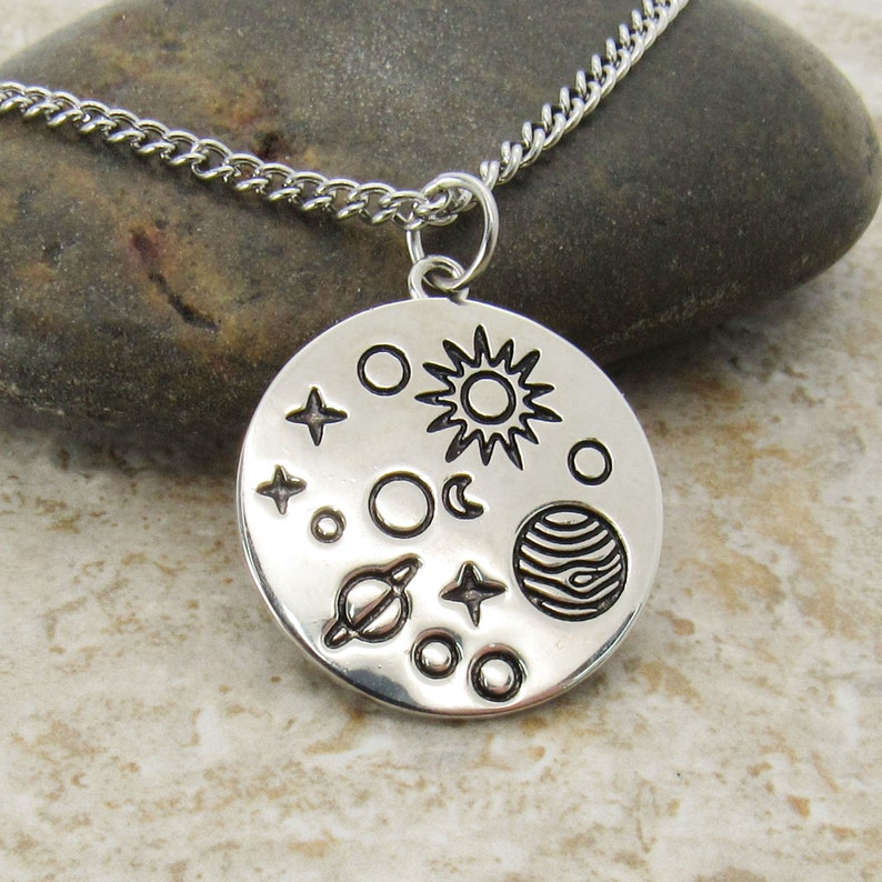 May include: A silver necklace with a round pendant featuring a stylized design of the sun, moon, stars, and planets.