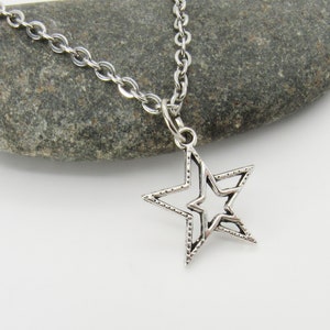 Puede incluir: Un collar de cadena de plata con un colgante en forma de estrella. La estrella tiene un doble contorno y una superficie texturizada.