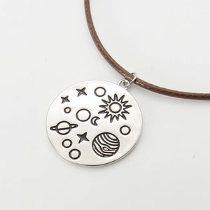 May include: A silver necklace with a round pendant featuring a sun, moon, stars, and planets. The pendant is attached to a brown leather cord.