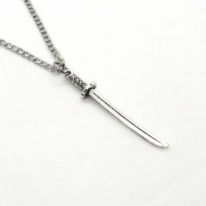 Puede incluir: Un collar de cadena de plata con un colgante de espada de plata. La espada tiene un mango detallado y una hoja curva.
