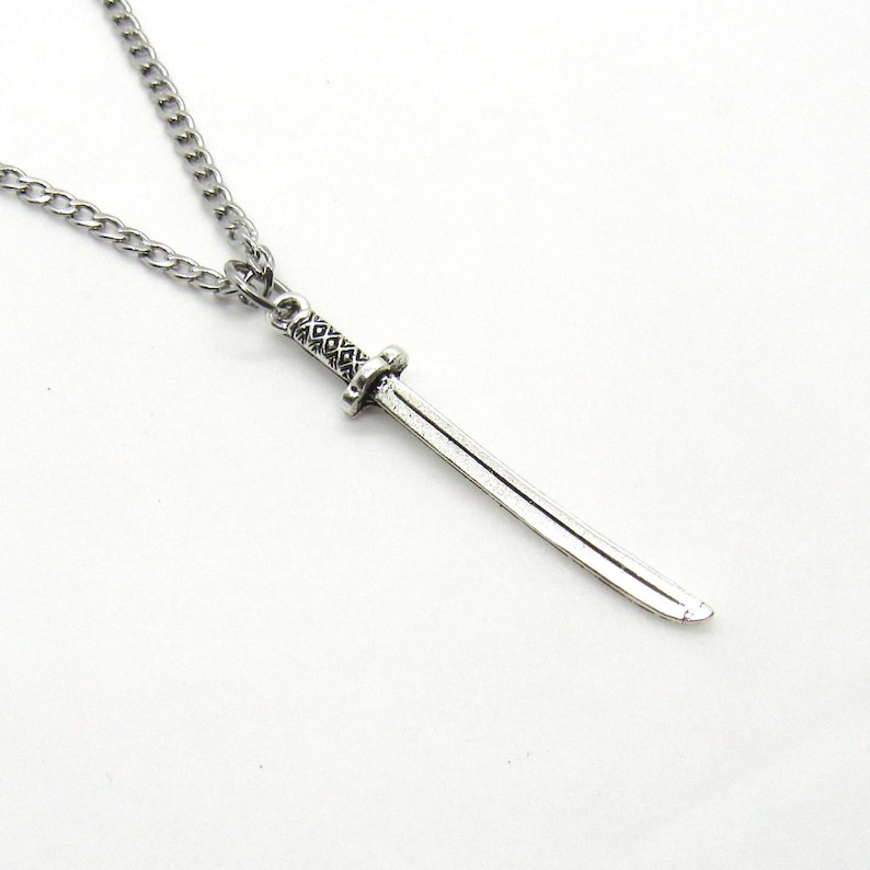 Puede incluir: Un collar de cadena de plata con un colgante de espada de plata. La espada tiene un mango detallado y una hoja curva.