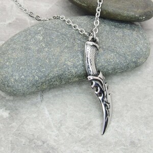 May include: A silver necklace with a detailed dagger pendant. The pendant has a textured handle and a sharp blade with intricate designs.