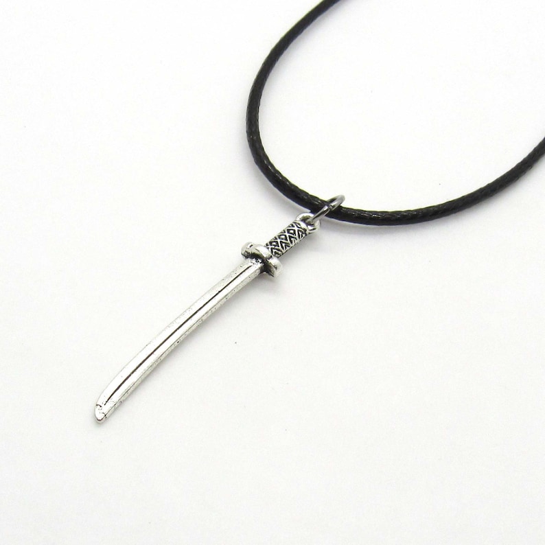 Puede incluir: Un collar con un colgante de espada de plata en un cord&oacute;n negro. La espada tiene un mango detallado y una hoja afilada.