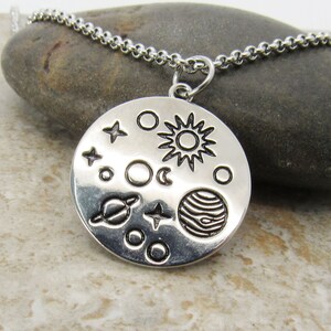 May include: A silver necklace with a round pendant featuring a stylized depiction of the solar system, including the sun, moon, planets, and stars.