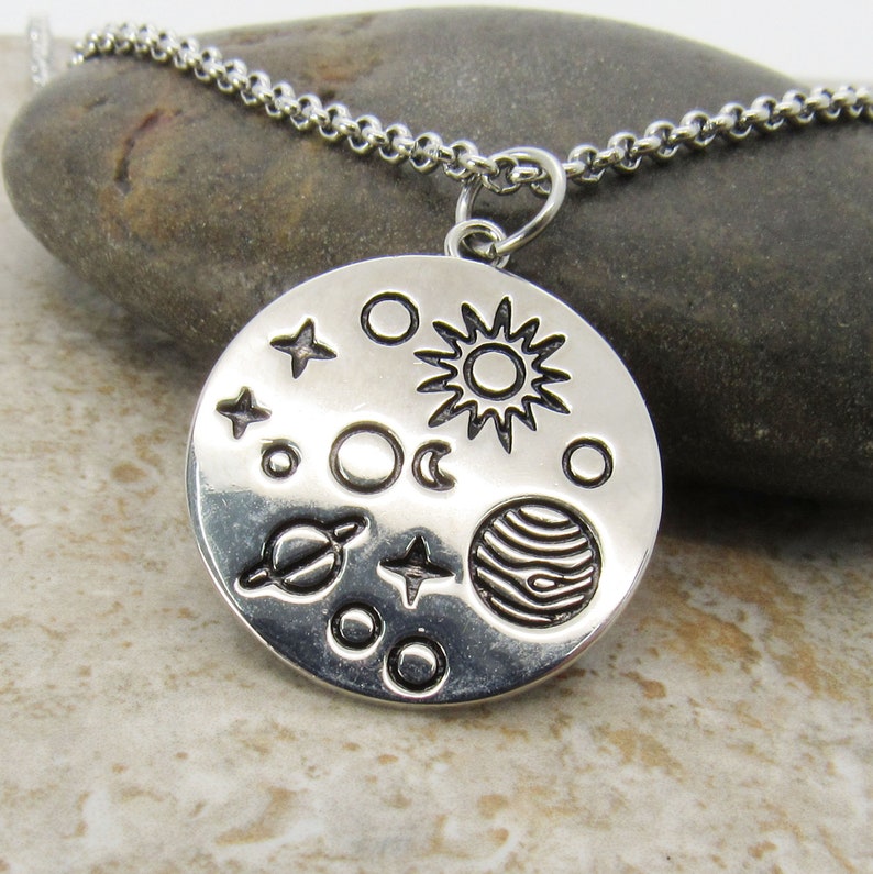 May include: A silver necklace with a round pendant featuring a stylized depiction of the solar system, including the sun, moon, planets, and stars.