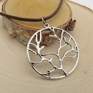 Op de afbeelding: Een zilveren ketting met een ronde hanger met een boom des levens ontwerp. De hanger is gemaakt van metaal en heeft een delicaat, ingewikkeld ontwerp. De ketting is aan een bruin leren koord.