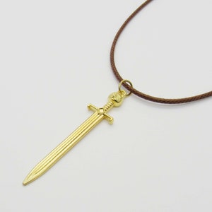 Pode incluir: Um colar com um pingente de espada dourada em um cord&atilde;o marrom. A espada tem uma empunhadura detalhada e uma l&acirc;mina longa e reta.