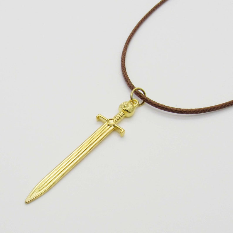 Pode incluir: Um colar com um pingente de espada dourada em um cord&atilde;o marrom. A espada tem uma empunhadura detalhada e uma l&acirc;mina longa e reta.