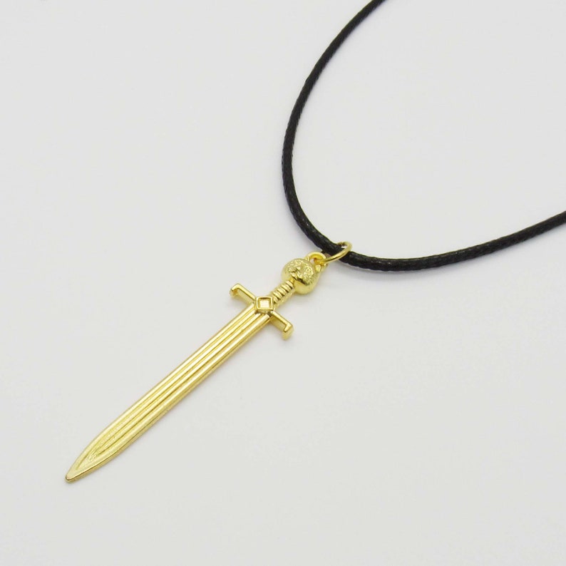 Pode incluir: Um colar com um pingente de espada dourada em um cord&atilde;o preto. A espada tem um design detalhado e uma pomo arredondada.