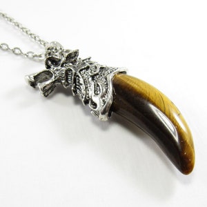 May include: A silver-colored wolf head pendant necklace with a large, curved, tiger's eye gemstone. The pendant is suspended from a delicate chain. The gemstone is a warm brown color with dark brown streaks.