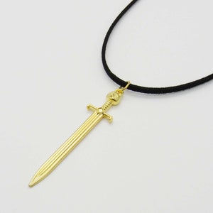 Pode incluir: Um colar com um pingente de espada dourada em um cord&atilde;o preto. A espada tem uma empunhadura detalhada e uma ponta afiada.