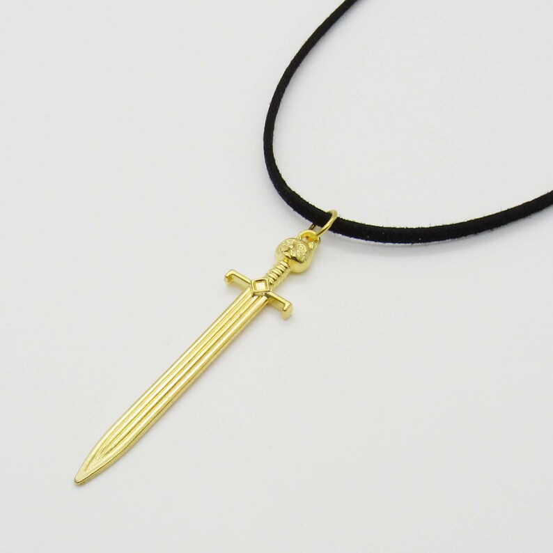Pode incluir: Um colar com um pingente de espada dourada em um cord&atilde;o preto. A espada tem uma empunhadura detalhada e uma ponta afiada.