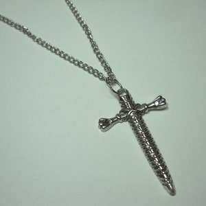 May include: A silver chain necklace with a sword-shaped pendant. The pendant is detailed with a braided design and has a cross shape.