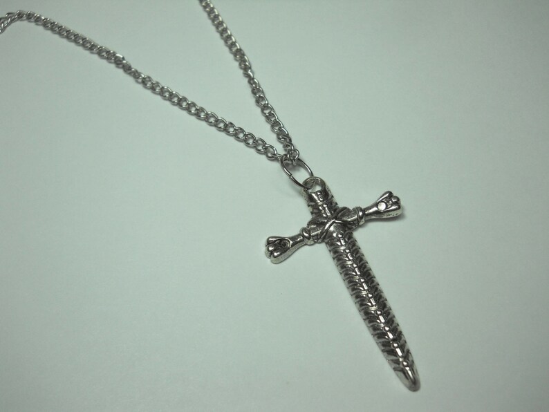 May include: A silver chain necklace with a sword-shaped pendant. The pendant is detailed with a braided design and has a cross shape.
