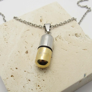 Op de afbeelding: Een ketting met een zilverkleurige ketting en een pilvormige hanger. De hanger is tweekleurig, met een zilveren bovenkant en een goudkleurige onderkant. Een kruis is geëtst op het gouden gedeelte. De tekst "be Mood" is geëtst op het zilveren gedeelte.