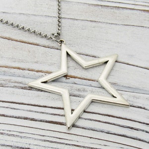 Puede incluir: Un collar con un colgante en forma de estrella de plata con un diseño simple y abierto. La cadena es de plata y tiene un aspecto delicado y enlazado.