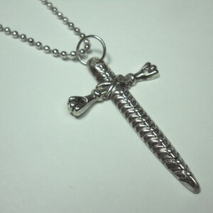 May include: A silver chain necklace with a sword-shaped cross pendant. The cross is detailed with a braided design and has a knotted detail at the center.