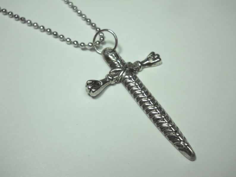 May include: A silver chain necklace with a sword-shaped cross pendant. The cross is detailed with a braided design and has a knotted detail at the center.