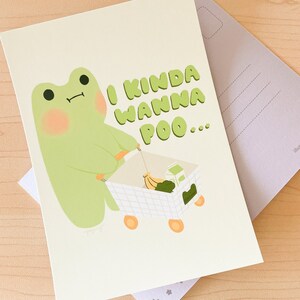 May include: A green frog with a pink blush on its cheeks pushes a miniature shopping cart filled with bananas and a carton of milk. The text "I kinda wanna poo..." is written in a bubble above the frog.