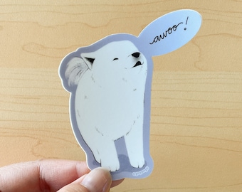 May include: A white cartoon dog sticker with a light blue speech bubble that says "awoo!". The dog is standing and howling. The sticker has a light purple border and is held by a hand.