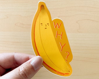 以下が含まれることがあります： 黄色いバナナのステッカーに、漫画の顔と「WHY?」の文字が書かれています。ステッカーは明るい木製の背景に貼られています。