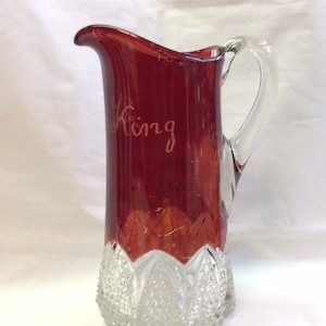 May include: A red glass pitcher with a clear glass handle. The pitcher has a clear glass base with a cut glass design. The pitcher has the word "King" etched on the side.