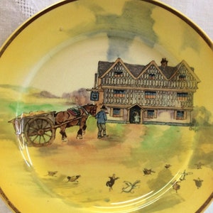 May include: A yellow ceramic plate with a gold rim features a watercolor painting of a Tudor-style house with a horse-drawn cart in front. The scene is set in a rural landscape with birds flying in the sky.
