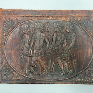 May include: A vintage, rectangular, brown leather book cover with an oval relief carving of figures in classical attire. The figures are arranged in a circle, holding hands. The border features ornate floral designs.