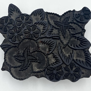 Könnte beinhalten: Ein dunkelgrauer, rechteckiger Holzblock mit einem Blumen- und Blättermuster. Das Design zeigt verschiedene Blumen- und Blattformen, die in die Oberfläche geschnitzt sind. Der Block wird wahrscheinlich zum Drucken oder Stempeln verwendet.