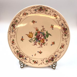 May include: A white ceramic plate with a gold rim and floral design. The plate features a bouquet of pink, yellow, and purple flowers with green leaves. The plate is on a gold metal stand.