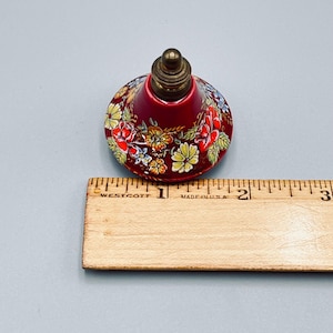 May include: A conical bottle stopper with a deep red base featuring a colorful floral pattern of yellow, orange, and red flowers. The top is a metallic brass-colored finial. A wooden ruler is placed alongside, showing a diameter of 1.75 inches.