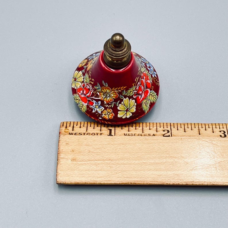 May include: A conical bottle stopper with a deep red base featuring a colorful floral pattern of yellow, orange, and red flowers. The top is a metallic brass-colored finial. A wooden ruler is placed alongside, showing a diameter of 1.75 inches.