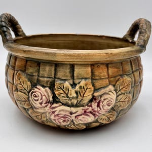 May include: A ceramic basket-shaped planter with two rope-style handles. The planter features a woven basket design with embossed roses and leaves in shades of pink, green, and brown. The interior is a darker brown.
