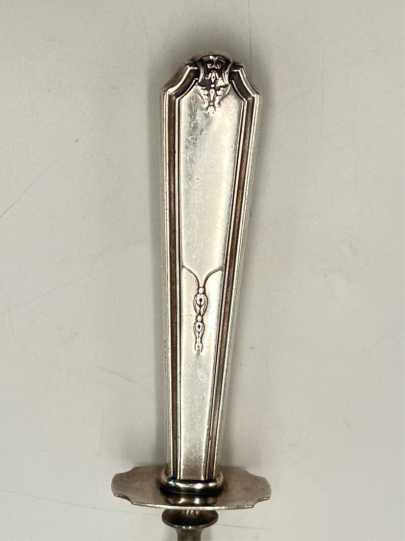 May include: Silver serving fork with an ornate handle and a decorative design. The handle is silver and has a floral design at the top. The fork has a long, thin blade.
