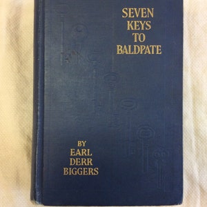 Puede incluir: Un libro de tapa dura azul con el título "Seven Keys to Baldpate" en letras doradas. El autor, Earl Derr Biggers, también está impreso en letras doradas en la portada.