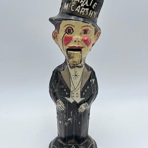 May include: Vintage Charlie McCarthy mechanical bank. The bank features a painted metal figure of a man in a tuxedo and top hat. The top hat is inscribed with the name "Charlie McCarthy". The figure has a hinged mouth and a coin slot.