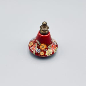 May include: A bell-shaped ceramic object with a glossy red finish, decorated with a colorful floral pattern featuring orange, yellow, and blue flowers. The top is capped with a metallic, tiered finial in a muted brass tone.